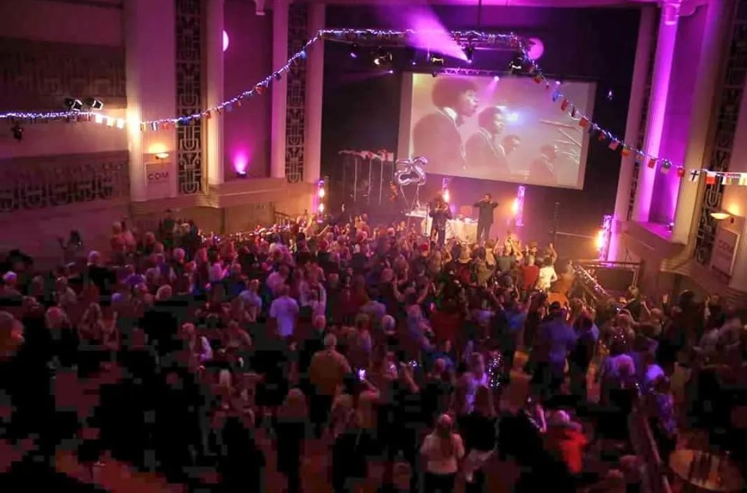 A packed dancefloor with raised hands, a stage and purple ballroom lighting